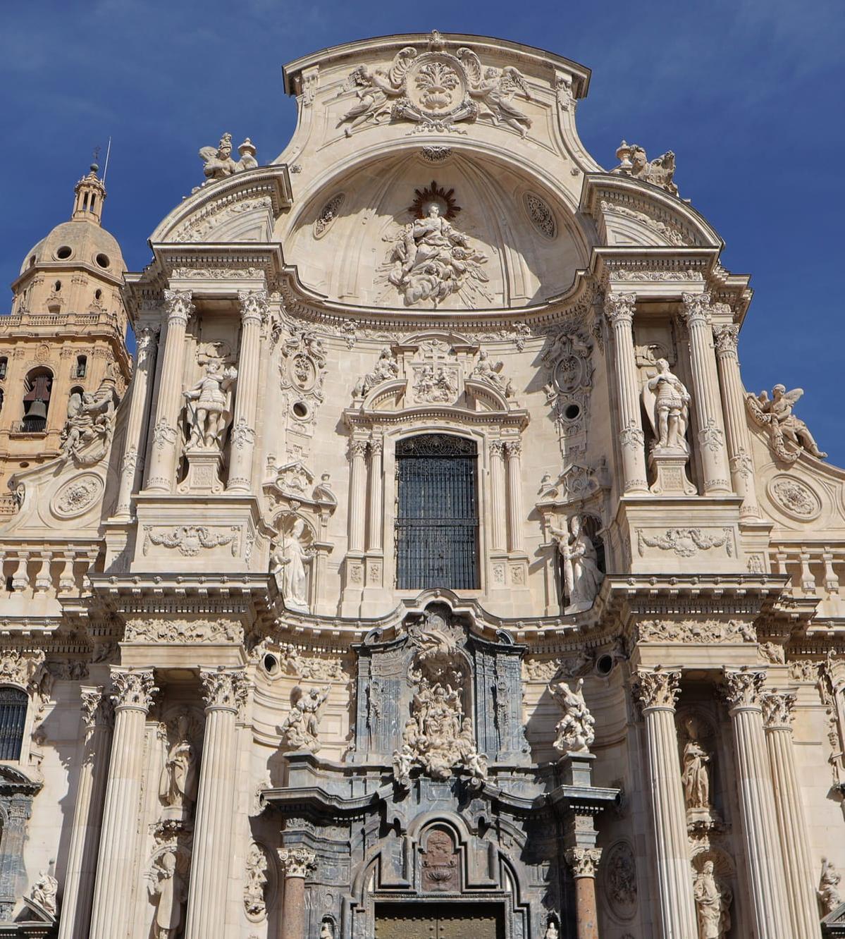 The Baroque main façade of Murcia Cathedral (Catedral de Santa María), carved in pale stone with statues and columns, part of the bell tower on the left, under a deep blue sky