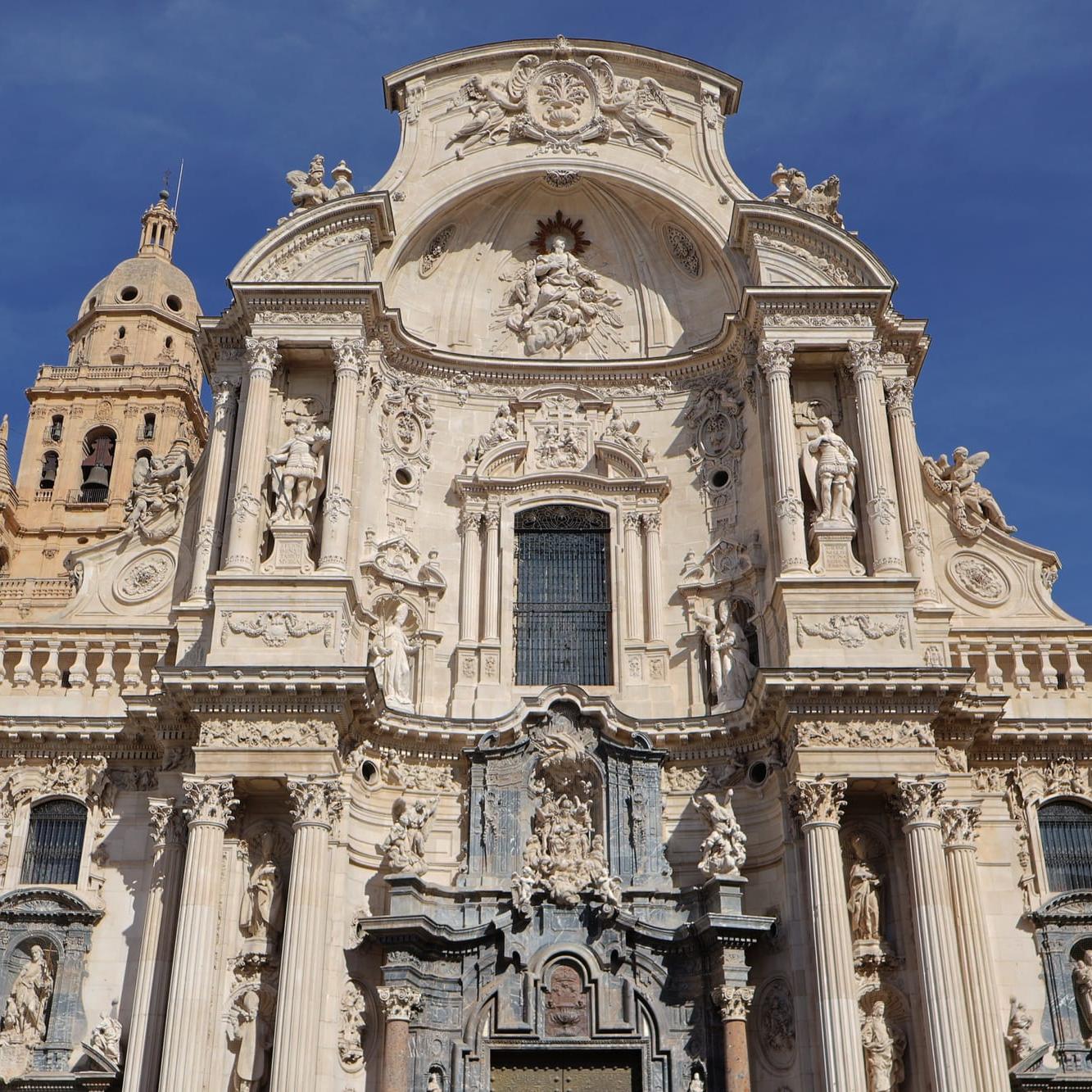 The Baroque main façade of Murcia Cathedral (Catedral de Santa María), carved in pale stone with statues and columns, part of the bell tower on the left, under a deep blue sky