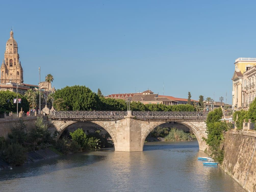 The river Segura flowing between stone embankments under the arched Puente de los Peligros (Old Bridge), with the cathedral bell tower rising over the trees of the city centre