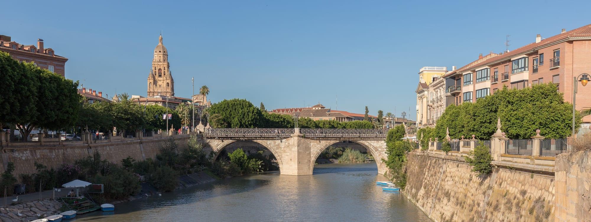 The river Segura flowing between stone embankments under the arched Puente de los Peligros (Old Bridge), with the cathedral bell tower rising over the trees of the city centre